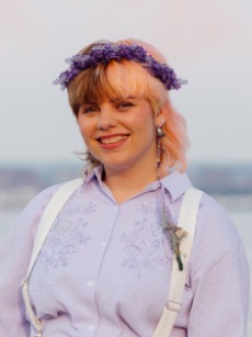 A portrait of a person with split dyed brown and pink hair wearing a purple flower crown and purple dress shirt.